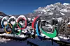 Olympiske ringer og Paralympiske symboler står ute i snøen. Bak dem ligger en fjellby med hus, trær og snødekte fjell i Italia. Det er klar blå himmel og vinterstemning.