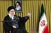 Irans øverste leder Ali Khamenei sitter på en stol og holder en tale. Han har svart turban, briller og langt, grått skjegg. Han peker med høyre hånd mens han snakker i to mikrofoner. Bak ham henger et portrett på veggen og et iransk flagg står til høyre.
