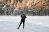 En person går på skøyter på isen på en frossen innsjø. Personen har mørke vinterklær, lue og ryggsekk, og bruker staver for å holde balansen. I bakgrunnen er det snødekte trær i lav vintersol.
