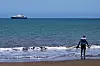 Cruiseskip ligger for anker ute på havet, med en person stående i vannkanten på stranden i forgrunnen.