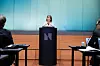 En kvinne står ved en talerstol foran en blå vegg i en pressekonferansesal med publikum ved bord på hver side.