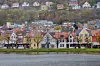 To busser kjører langs en gate ved vannet, med tett bebyggelse og åsside i bakgrunnen.