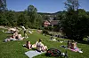 Mange mennesker ligger på tepper og bader på en grønn badeplass ved elva, med trær og bygninger i bakgrunnen.