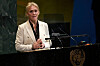 En kvinne i lys blazer taler ved talerstolen i FNs sal med FN-emblemet foran.