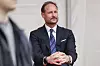 En mann i mørk dress og blått slips sitter med hendene foldet foran seg under et arrangement.