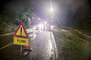 Et gult flomskilt står ved en vannfylt vei i mørket, med lys fra biler i det fjerne.