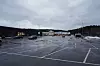 Stor, nesten tom parkeringsplass ved et kjøpesenter, med noen biler, våt asfalt og overskyet himmel.