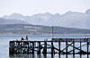Mennesker står og går på en brygge ved vannet, med fjell i bakgrunnen og klart sommerlys.