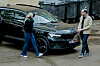 En svart bil står parkert utenfor et bygg mens to personer står ved siden av den på en grå asfaltert plass.