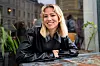 En smilende kvinne sitter ved et bord på en kafé med planter og bygninger i bakgrunnen.