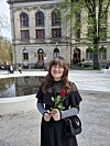 En person med briller holder en rød rose foran en stor steinbygning og et grunt vannspeil.