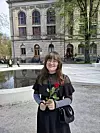 En person med briller holder en rød rose foran en stor steinbygning og et grunt vannspeil.