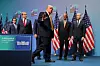 Donald Trump går fra et podie på en scene med flere menn i dress og amerikanske flagg i bakgrunnen.