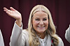 Kronprinsesse Mette-Marit smiler og vinket med løftet hånd foran et mørkerødt bakteppe.