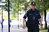 En politibetjent går langs en fortauvei i et bymiljø med trær og skilt i bakgrunnen.