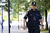 En politibetjent går langs en fortauvei i et bymiljø med trær og skilt i bakgrunnen.