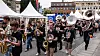 Et korps med saksofoner, tromboner og sousafoner marsjerer i en gate omgitt av publikum og telt.