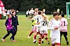 Ungdomsspillere i fotballdrakter står og løper på en gressbane med tilskuere og trenere i bakgrunnen.