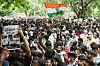 Folkemengde protesterer i New Delhi; en demonstrant holder en hindi-plakat, mens et indisk flagg vaier over mengden.