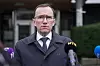 En mann i mørk frakk står foran flere mikrofoner utenfor en bygning.