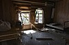 Et mørkt, skadet rom med ødelagte møbler, knust materiale på gulvet og skader rundt vinduene.