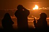 Silhuetter av personer som ser en delvis solformørkelse over havet ved solnedgang.