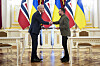 Jonas Gahr Støre og Volodymyr Zelenskyj tar hverandre i hendene foran norske og ukrainske flagg i en sal i Kyiv. Begge holder hver sin utgave av avtalen de har signert.