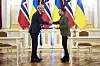 Jonas Gahr Støre og Volodymyr Zelenskyj tar hverandre i hendene foran norske og ukrainske flagg i en sal i Kyiv. Begge holder hver sin utgave av avtalen de har signert.