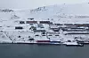 Luftfoto av Barentsburg med bygninger, kai og snødekte fjellsider ved sjøen.