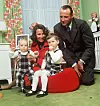 Kronprinsfamilien sitter samlet i en stue med grønne tepper og blomstrete gardiner.