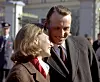 Kong Harald og dronning Sonja smiler på Slottsplassen foran Slottet mens folk og vakter ses i bakgrunnen.