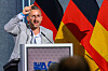Mann ved talerstol med AfD-logo, foran flere tyske flagg under en politisk tale.