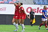 To Brann-spillere i røde drakter feirer på banen mens en Vålerenga-spiller står i bakgrunnen.