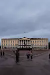 Slottet i Oslo med grå himmel og folk i forgrunnen, sett fra Slottsplassen under forberedelsene til gravferden.