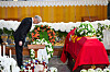 En mann i mørk dress bøyer seg foran en kiste dekket med rødt klede og blomster i en kirke.