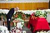 En mann i mørk dress bøyer seg foran en kiste dekket med rødt klede og blomster i en kirke.