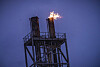 Industriell fakkel på toppen av en metallplattform med flammer mot blå kveldshimmel.