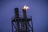 Industriell fakkel på toppen av en metallplattform med flammer mot blå kveldshimmel.