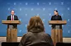 To politikere står ved hver sin talerstol foran blå bakgrunn, sett fra ryggen til en person i forgrunnen.
