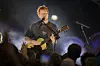 Ed Sheeran står på en opplyst scene med gitar, mens publikum rekker hendene i været foran ham.