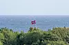 Norsk flagg på en flaggstang over grønn kystvegetasjon med havet bak.