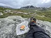 Matpause på en stein på Hardangervidda med fjellsko, turbukse og matpakket inn i DNT-papir.