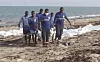 Kystvakter bærer omkomne migranter på en sandstrand, mens hvite likposer ligger langs vannkanten.