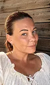 Nærbilde av en kvinne med oppsatt hår og hvit bluse foran en trevegg.