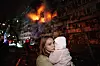 En ung kvinne holder et lite barn foran et brennende, skadet boligbygg i Kyiv.