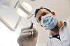 Maskert tannlege i hvit frakk holder et dentalinstrument under en tannlegelampe.