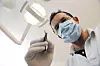 Maskert tannlege i hvit frakk holder et dentalinstrument under en tannlegelampe.