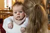 Baby med smekke holdes på armen av moren og ser mot kameraet i et lyst hjem.