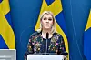 Bildet er av Sveriges sosialminister Lena Hallengren. Hun står foran tre svenske flagg og en blå vegg. Hun snakker i en mikrofon som er festet i en talerstol.