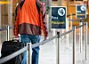 Bildet er av ryggen til en person med trillekoffert som går langs et sperrebånd. Han passerer et skilt hvor det står at han nærmer seg passkontroll.